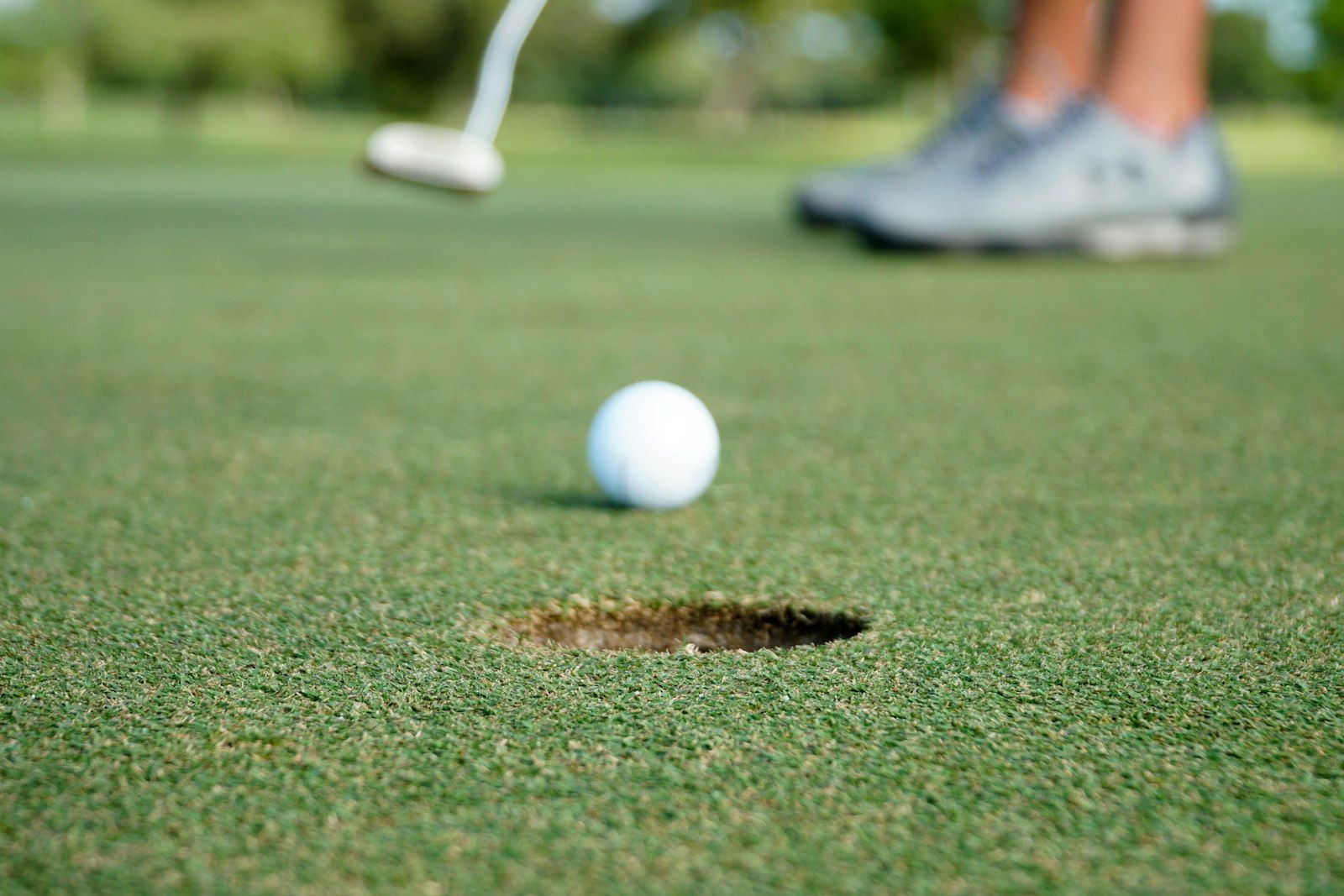 Balle de golf près du trou sur un green, avec un putter et des pieds flous à l'arrière-plan